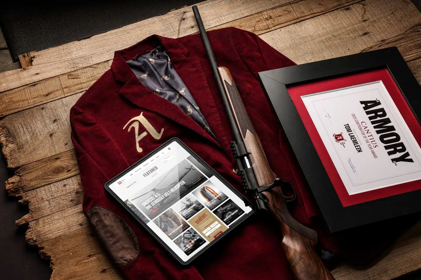 A framed Cantius Award certificate presented to Tom Laemlein, shown beside the crimson Cantius Award jacket with its gold embroidered A, a rifle, and a tablet displaying The Armory Life website.